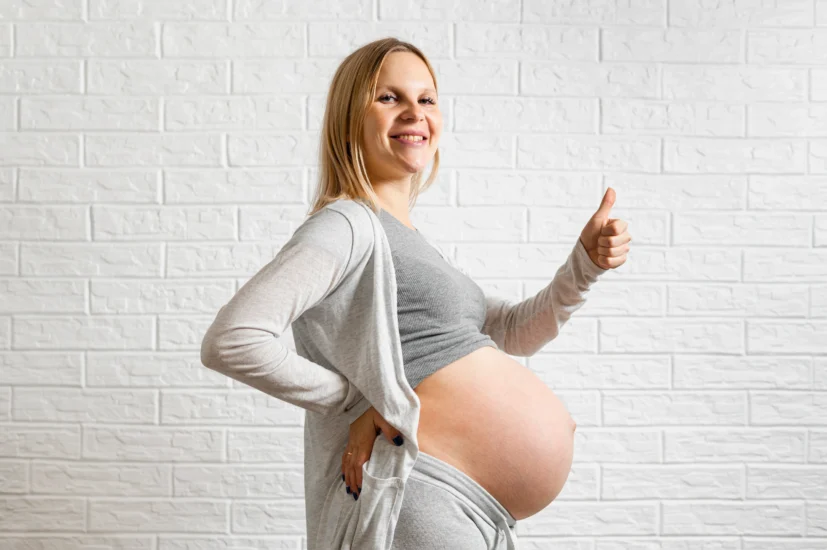 Kobieta w zaawansowanej ciąży, która jest w trakcie przygotowanie do porodu, stoi bokiem i unosi kciuk.