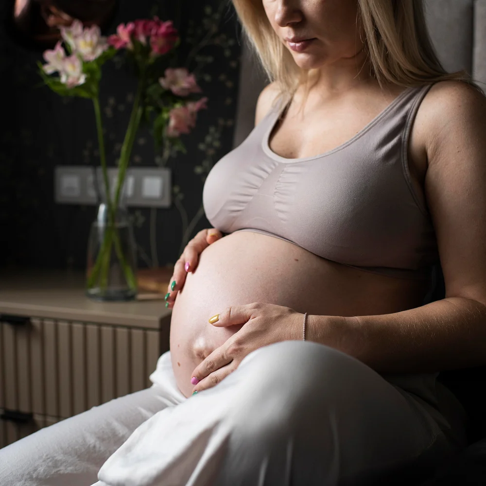 Kobieta w beżowym topie w pokoju z kwiatami trzyma brzuch podczas, w którym rozwija się jej ciąża tydzień po tygodniu.