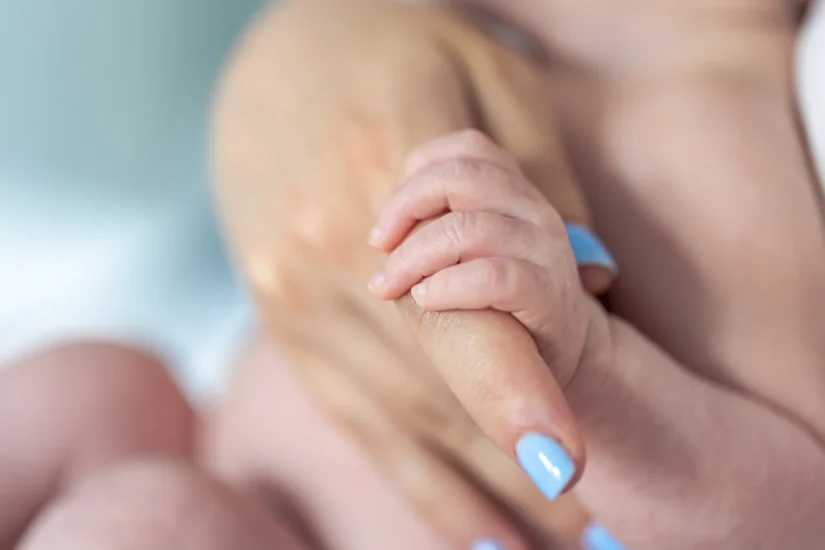 Noworodek trzyma palcem dłoń kobiety po porodzie, bliski kontakt matki z dzieckiem w okresie połogu, wsparcie emocjonalne po porodzie i szkoła rodzenia.